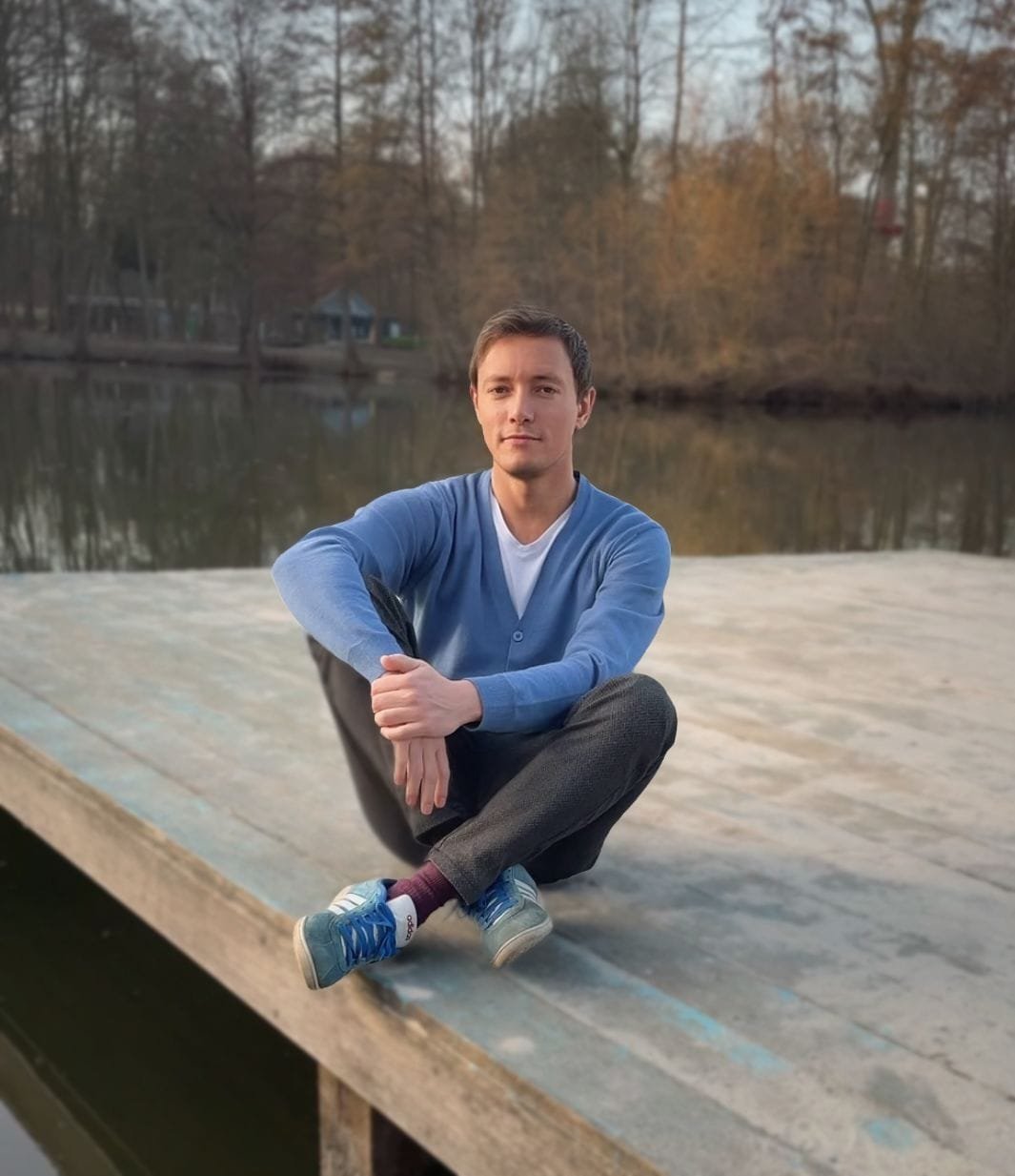 Ein junger Mann in einer blauen Strickjacke sitzt auf einem Holzsteg am Ufer eines Sees, umgeben von kahlen Bäumen in einer ruhigen Naturlandschaft.